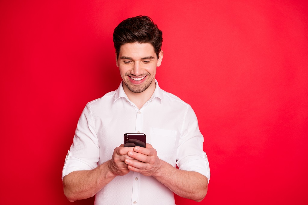 Texas Defensive Driving photo of smiling modern man typing message to someone with his telephone while isolated with red background